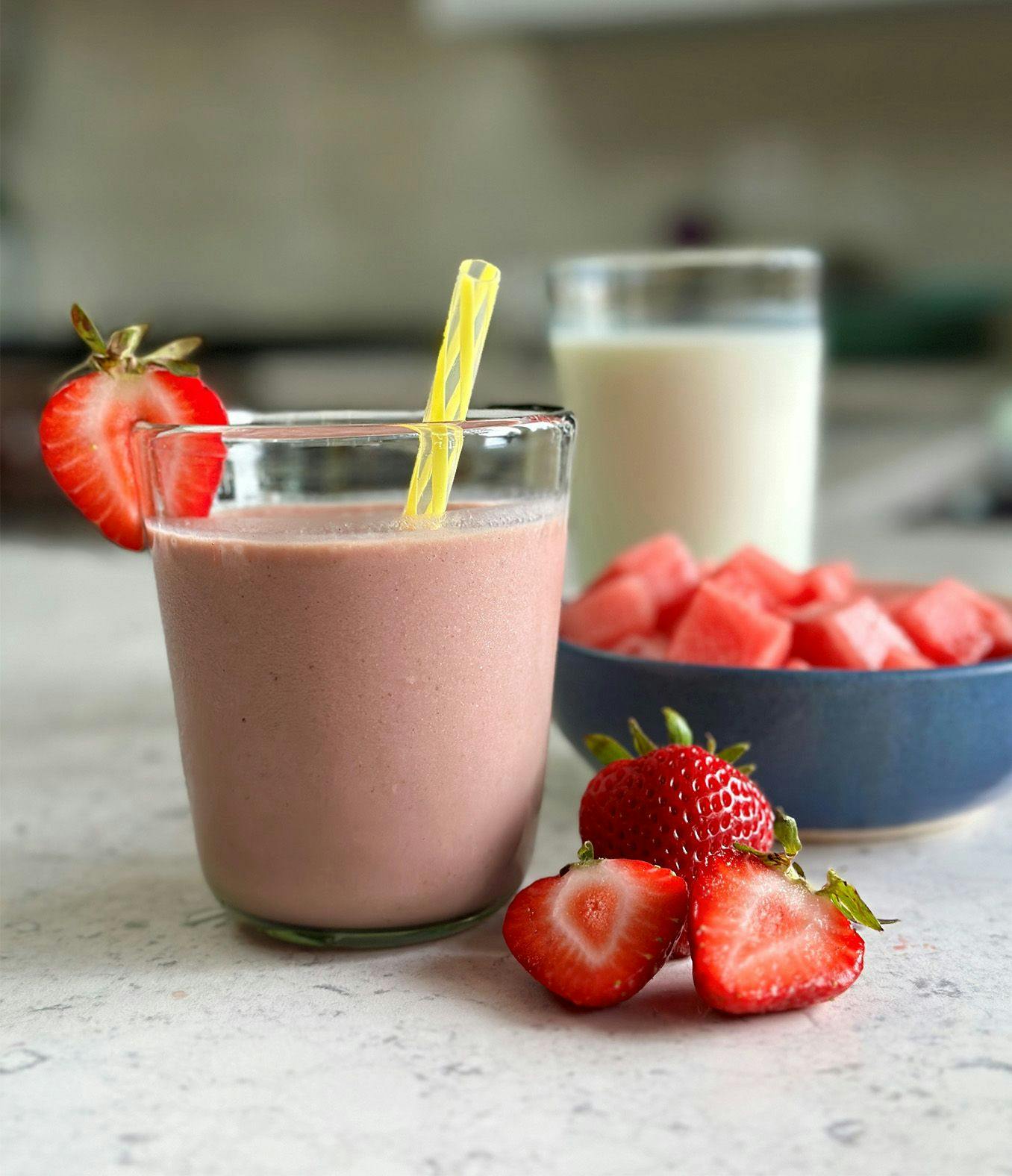 Smoothie with a yellow straw and a strawberry garnish sitting next to a bowl of watermelon. 