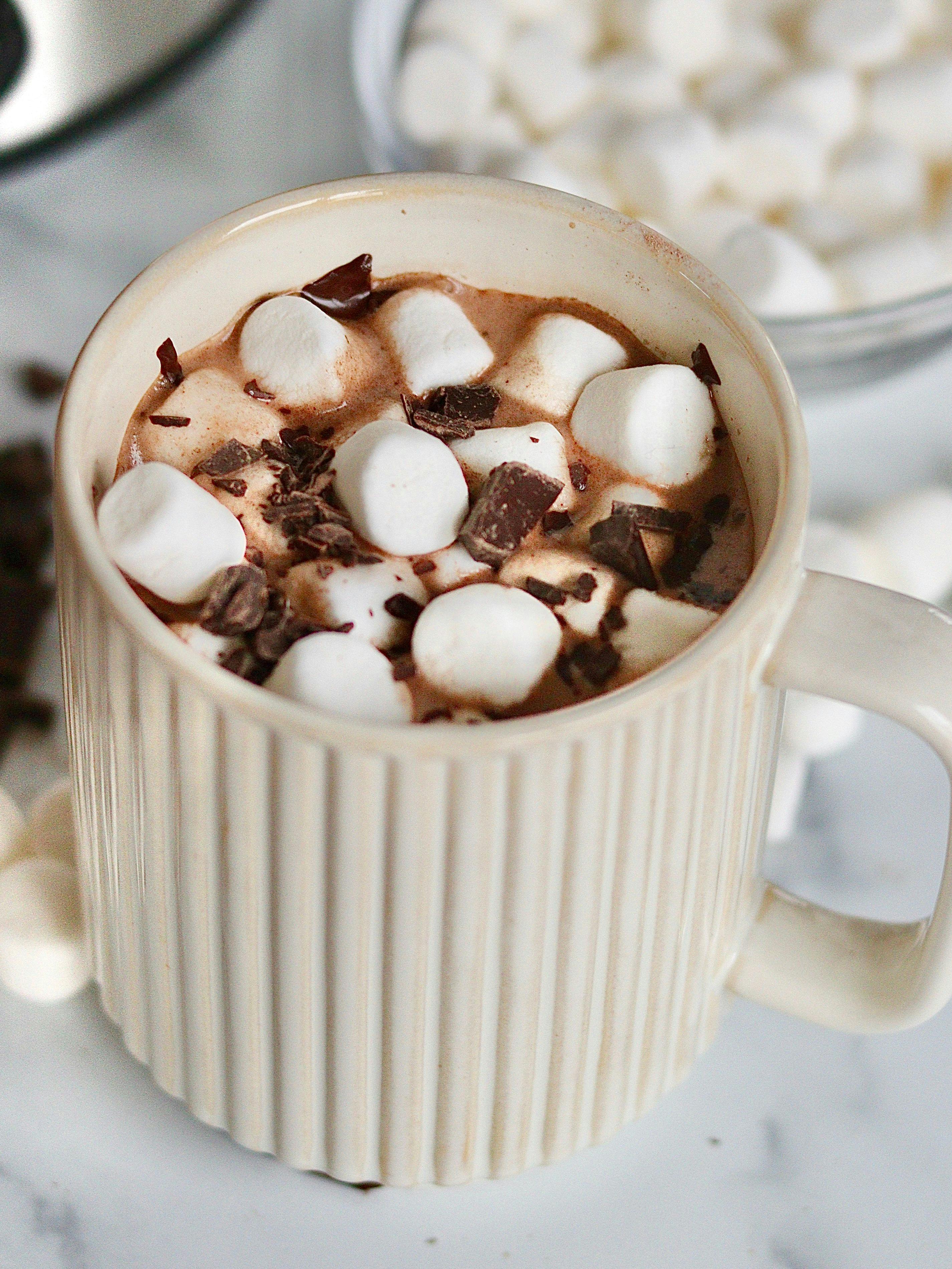 A mug of hot chocolate next to a slow cooker, chocolate shavings, marshmallows and organic dairy products used to make hot chocolate.