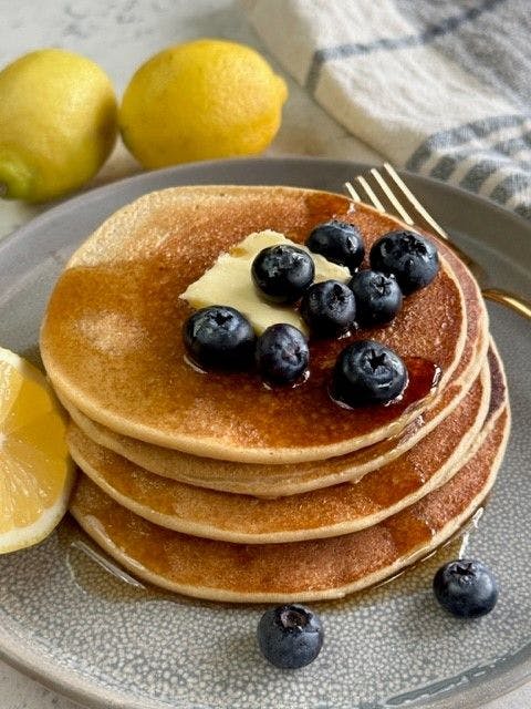 a stack of Blender Pancakes with blueberries and butter on top and they are on a plate with lemons and a fork next to them.
