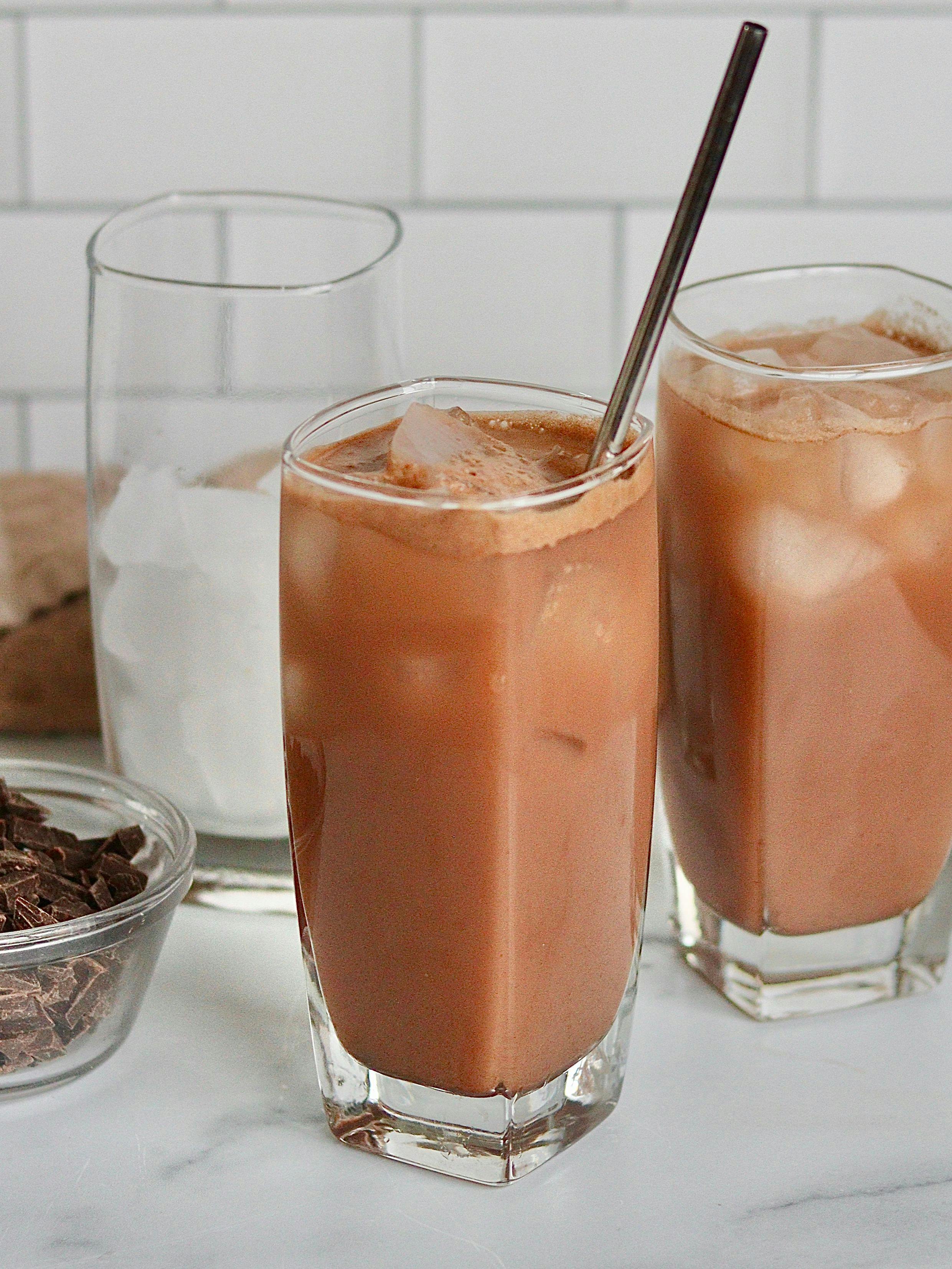 Two glasses of iced hot chocolate on a counter with organic whole milk and chocolate pieces.